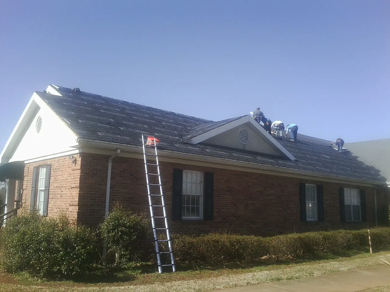 Crew tearing shingles off a brick ranch roof with a ladder set — Metal Roof Repair work by our crew in Fletcher
