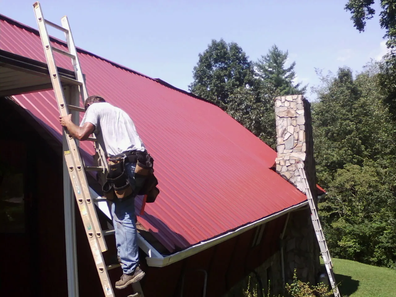 Roofer on a ladder beside a red metal roof and stone chimney — Soffit & Fascia Repair by Roofing Brothers in Fletcher